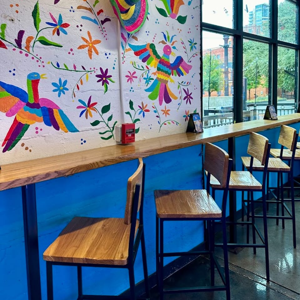 Mas Street Tacos interior with colorful mural, wooden bar, and stools, overlooking a city street.