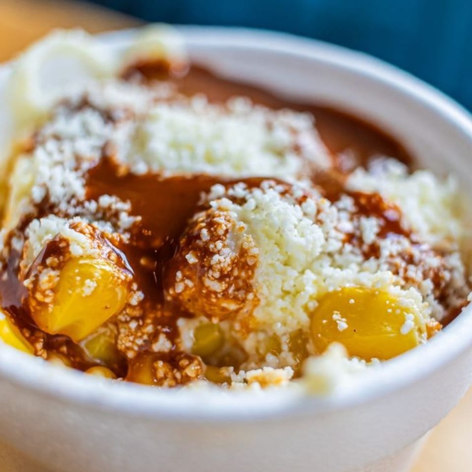 Close-up of Mas Street Tacos' esquites, featuring corn, cotija cheese, and chili sauce in a white bowl.