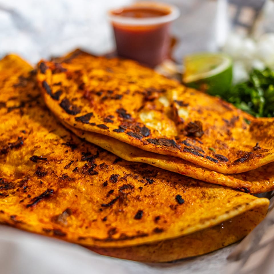 Close-up of three golden-brown birria tacos with a side of red sauce, lime, and cilantro.