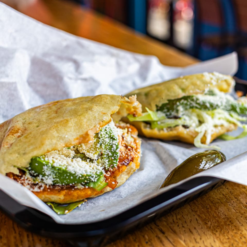 Two Mas Street Tacos, one open with avocado, cheese, and lettuce, served with a pickle.