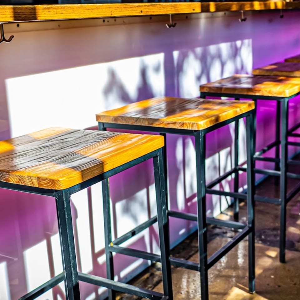 Mas Street Tacos bar area with wooden stools, purple lighting, and sunlit shadows on the wall.