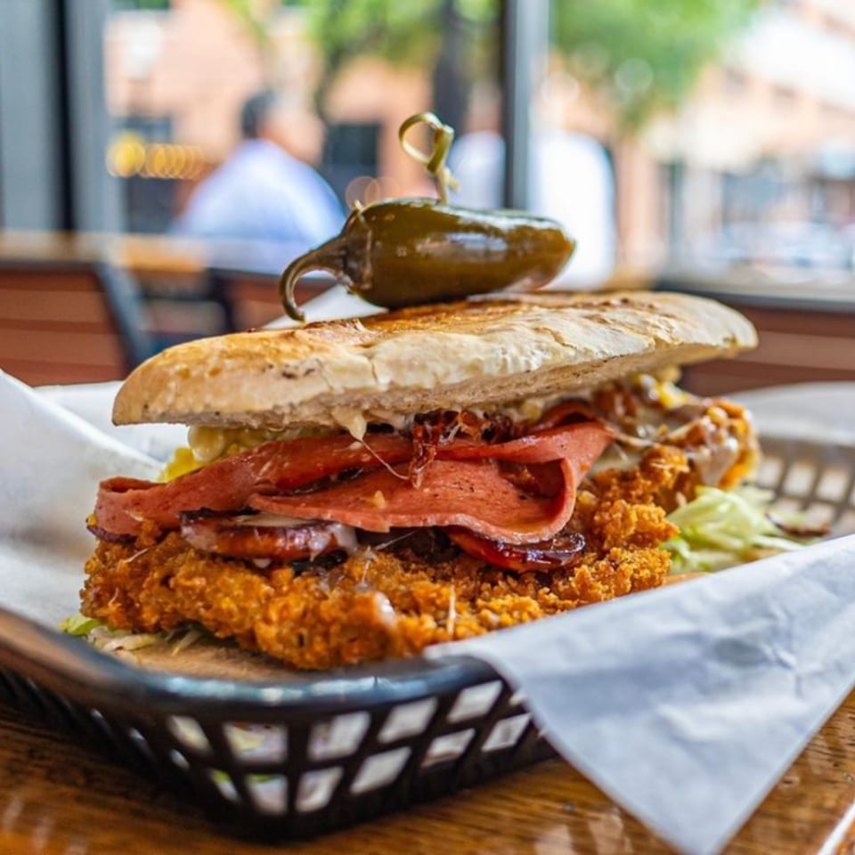 A large fried chicken sandwich with deli meat and a jalapeño on top, in a basket.